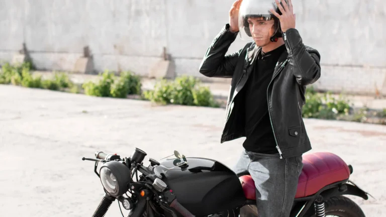 A man in a black leather jacket and jeans is putting on a clear helmet, standing next to a black cafe racer motorcycle with a red seat.
