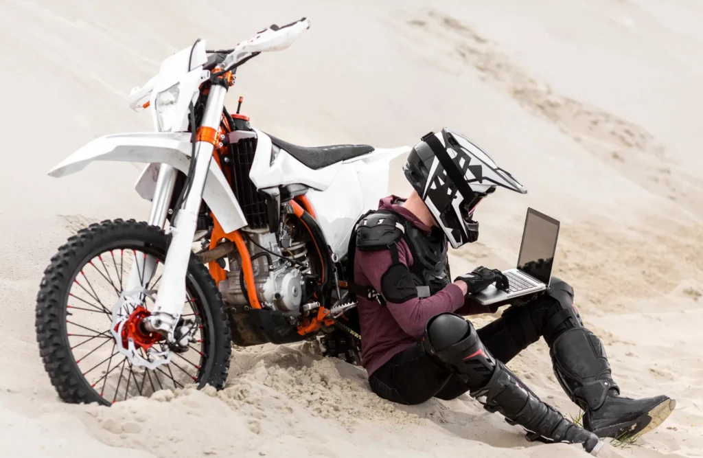 Dirt bike rider in motocross gear using laptop in desert, off-road motorcycle adventure with enduro bike.